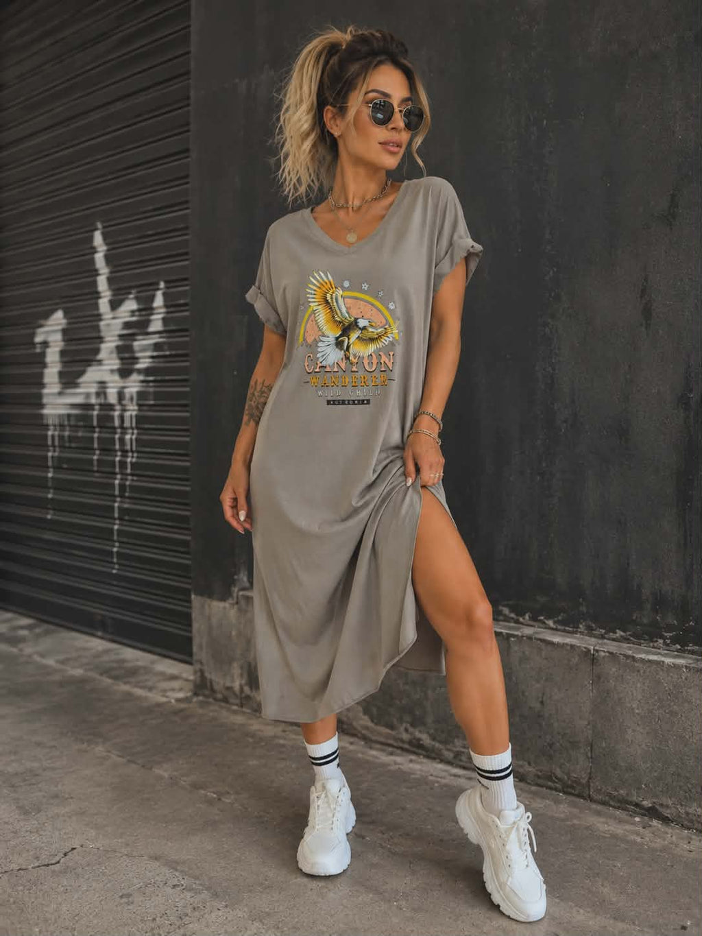 Damska sukienka oversize Canyon Wanderer w kolorze taupe z bawełny, z nadrukiem orła i rozcięciem po boku, zestawiona z białymi sneakersami w miejskiej stylizacji.