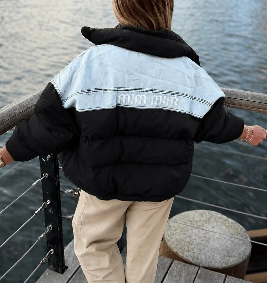 Tył czarnej damskiej kurtki puchowej oversize z jasnymi jeansowymi wstawkami. Pikowana kurtka Boska Lady prezentowana nad wodą.